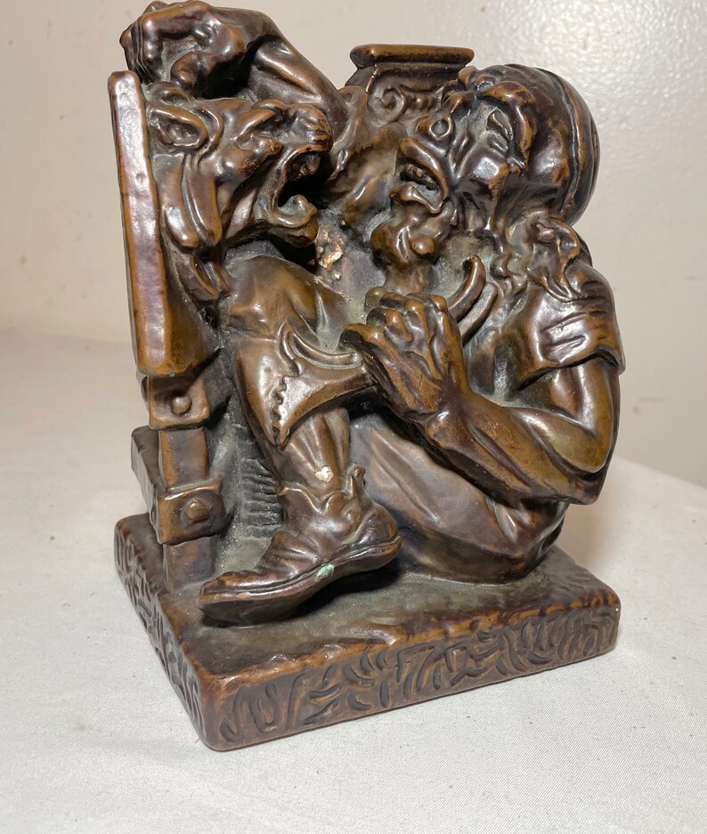 Puede incluir: Un sujetalibros de bronce que representa a un hombre siendo atacado por una g&aacute;rgola. El hombre lleva una t&uacute;nica y un turbante, y la g&aacute;rgola le est&aacute; ara&ntilde;ando la cara. El sujetalibros est&aacute; sobre una base cuadrada con un dise&ntilde;o decorativo.