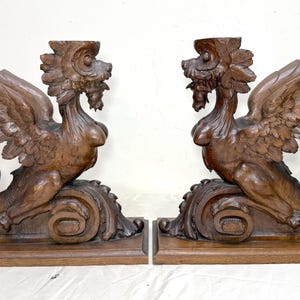 May include: Pair of intricately carved wooden griffin sculptures. Each sculpture features a winged creature with a lion's body, a human-like torso, and a bird's head. The wings are spread, and the tail curls. The sculptures are brown.