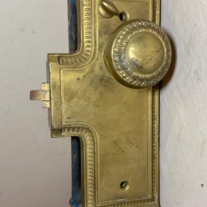 May include: Antique brass door lock with ornate detailing. The rectangular plate features a round doorknob and a lever. The lock has a vintage aesthetic, with signs of wear and tear, and is mounted on a blue painted surface.