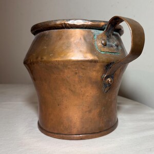 May include: A copper pitcher with a handle. The pitcher has a wide, rounded body that tapers towards the base. The handle is attached with rivets. The copper has a warm, reddish-brown color with some areas of green oxidation.