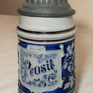 May include: A blue and white ceramic beer stein with a pewter lid. The stein features a floral design and the word "Prosit" in a decorative script.