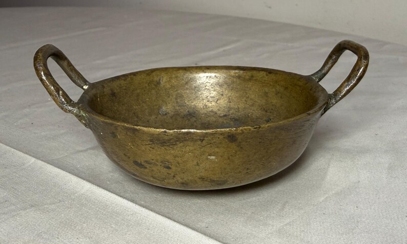 Puede incluir: Una taza de cer&aacute;mica blanca con asa negra y el texto "But First, Coffee" en negrita negra. La taza se encuentra sobre un fondo borroso, sugiriendo un ambiente acogedor.