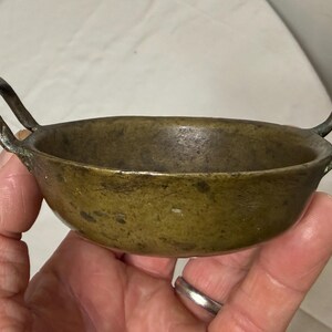 Puede incluir: Un peque&ntilde;o cuenco de bronce antiguo con dos asas de bucle. El cuenco tiene forma redondeada y una p&aacute;tina oscura y envejecida. Las asas est&aacute;n unidas a los lados del cuenco.