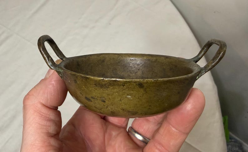 Puede incluir: Un peque&ntilde;o cuenco de bronce antiguo con dos asas de bucle. El cuenco tiene forma redondeada y una p&aacute;tina oscura y envejecida. Las asas est&aacute;n unidas a los lados del cuenco.