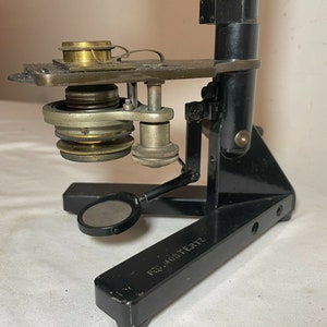 May include: An antique black microscope with brass and silver-colored components. The microscope has a base, a vertical support, and an angled arm holding the lens and stage. The base has the text "FRD. MOSTERTZ".