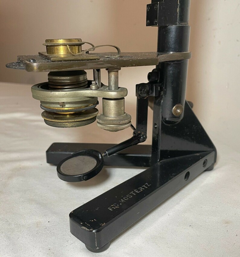 May include: An antique black microscope with brass and silver-colored components. The microscope has a base, a vertical support, and an angled arm holding the lens and stage. The base has the text "FRD. MOSTERTZ".