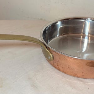May include: A copper-colored frying pan with a stainless steel interior and a brass handle. The pan is oval-shaped and has a long, curved handle. The handle is attached to the pan with a rivet. The pan is sitting on a white surface.