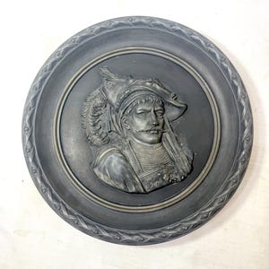 May include: A dark gray, round wall plaque featuring a bas-relief portrait of a man in a hat with a feather and a long mustache. The portrait is set within a circular frame with a decorative border.