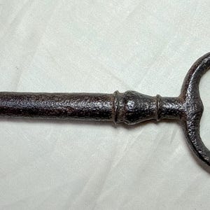 May include: A large, antique-style key with a dark, rusty brown finish. The key has a rectangular head, a long, cylindrical shaft, and a circular handle. The key is likely made of metal and appears to be aged.