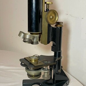 May include: An antique microscope with a black body and brass accents. The microscope has multiple lenses, adjustment knobs, and a stage for slides. The base is marked with text. A classic scientific instrument.