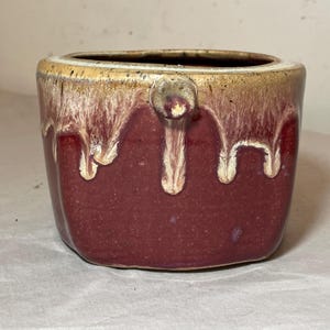 May include: A red and white ceramic planter with a unique glaze pattern. The planter has a rounded shape and a small handle on the side.