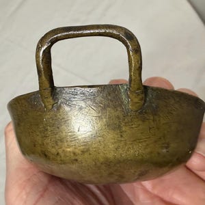 Puede incluir: Un peque&ntilde;o cuenco antiguo de bronce con asa. El cuenco tiene forma redondeada y una superficie ligeramente texturizada. El asa es arqueada y est&aacute; unida al borde del cuenco. El bronce tiene una p&aacute;tina oscura y envejecida.