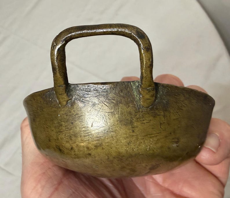 Puede incluir: Un peque&ntilde;o cuenco antiguo de bronce con asa. El cuenco tiene forma redondeada y una superficie ligeramente texturizada. El asa es arqueada y est&aacute; unida al borde del cuenco. El bronce tiene una p&aacute;tina oscura y envejecida.