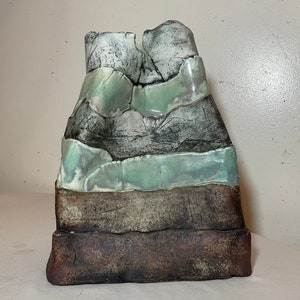 May include: A ceramic sculpture resembling a layered rock formation. The sculpture is made of multiple layers of clay, each with a different color and texture. The top layer is a light green, the middle layer is a dark brown, and the bottom layer is a light brown. The sculpture is a unique and interesting piece of art.