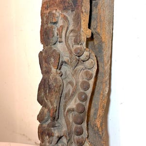 May include: A weathered, carved wooden architectural element. The dark brown wood features intricate relief carvings of figures and floral patterns. A series of round, raised details runs vertically along the side. The top has a rounded, decorative finial.