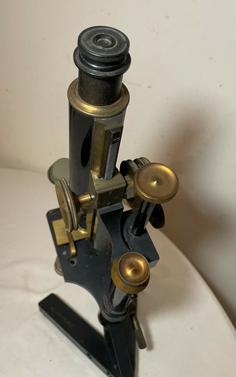 May include: An antique microscope with a black body and brass accents. The microscope has a cylindrical eyepiece, adjustment knobs, and a base. It is a vintage scientific instrument.