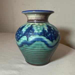 May include: A ceramic vase with a wide, rounded body and a narrow neck. The vase features a turquoise base with horizontal ridges, transitioning to a blue and turquoise patterned midsection. The rim is blue, with a brown and cream-colored band below.