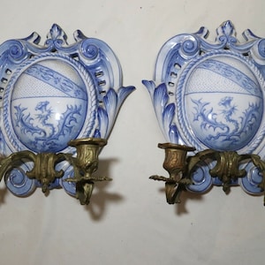 May include: Pair of antique wall sconces. Each sconce features a white and blue porcelain design with a central shield motif and a brass candle holder. The porcelain has a decorative floral and rope design. The sconces are mounted on a white wall.