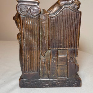 Puede incluir: Una escultura de bronce de una figura reclinada sobre una columna. La escultura est&aacute; detallada con intrincados tallados y tiene una superficie texturizada.