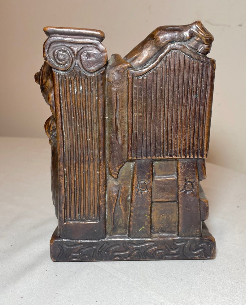 Puede incluir: Una escultura de bronce de una figura reclinada sobre una columna. La escultura est&aacute; detallada con intrincados tallados y tiene una superficie texturizada.