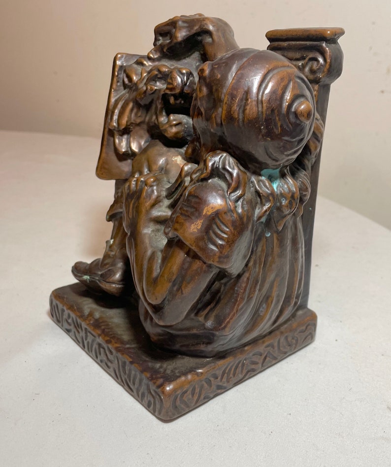 Puede incluir: Un par de sujetalibros de bronce que representan dos figuras, una sentada y la otra de pie, con detalles intrincados y una base texturizada.