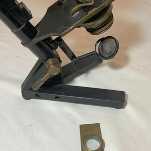 May include: An antique microscope with a black frame and brass accents. The microscope has a hinged arm, adjustable lenses, and a separate rectangular brass slide holder. The design suggests a vintage scientific instrument.