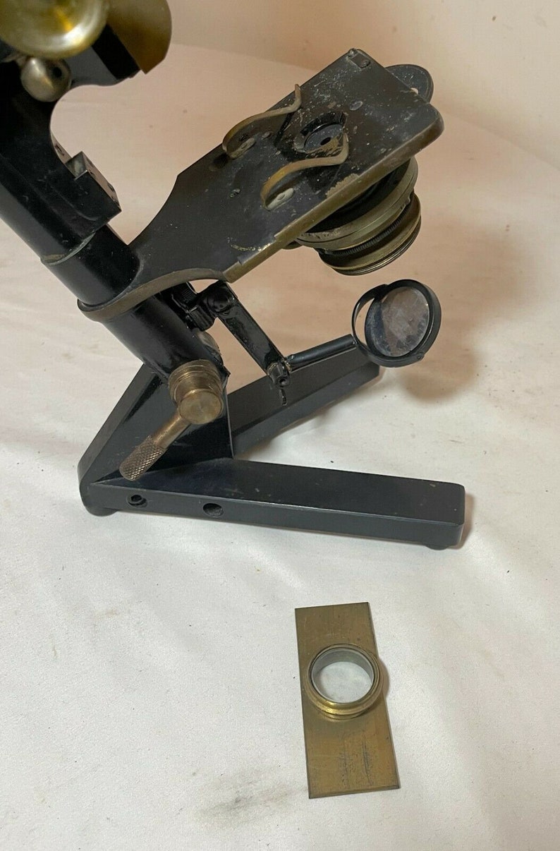 May include: An antique microscope with a black frame and brass accents. The microscope has a hinged arm, adjustable lenses, and a separate rectangular brass slide holder. The design suggests a vintage scientific instrument.