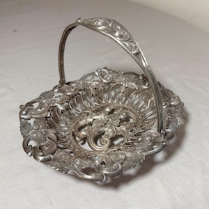 May include: Silver-toned metal basket with an ornate, openwork design. The basket features a handle and is decorated with floral and leaf motifs. The basket's shape is oval, and the handle is arched. The basket is likely vintage or antique.