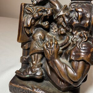 Puede incluir: Un sujetalibros de bronce que representa a un hombre luchando contra un le&oacute;n. El hombre lleva una toga y tiene barba. El le&oacute;n ruge y tiene las garras afuera. El sujetalibros es detallado y tiene una base texturizada.