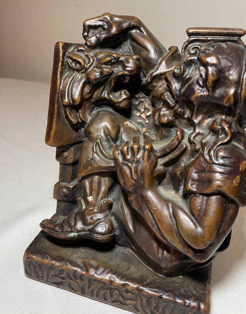 Puede incluir: Un sujetalibros de bronce que representa a un hombre luchando contra un le&oacute;n. El hombre lleva una toga y tiene barba. El le&oacute;n ruge y tiene las garras afuera. El sujetalibros es detallado y tiene una base texturizada.