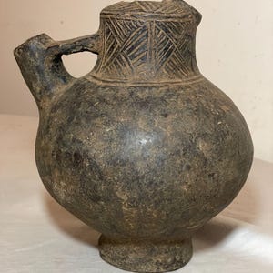 May include: A dark brown ceramic pitcher with a rounded body and a short spout. The pitcher has a handle and a base. The pitcher is decorated with a geometric pattern around the top.