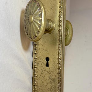 May include: Antique brass door hardware set. The set includes a rectangular backplate with ornate detailing, a keyhole, and two round doorknobs. The hardware has a vintage, aged appearance.
