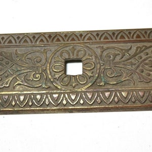 May include: A long, oval-shaped, brass plate with an intricate, embossed design. The plate has a central square hole and is decorated with repeating floral motifs.