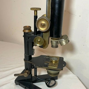 May include: An antique microscope with a black body and brass accents. It features adjustable knobs, a stage for slides, and a lens system. The microscope is mounted on a black base and appears to be in good condition.