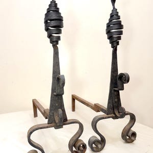 May include: Pair of antique black wrought iron fireplace andirons. Each andiron features a spiral design at the top, a decorative central support, and scrolled feet. The andirons are designed to hold logs in a fireplace.