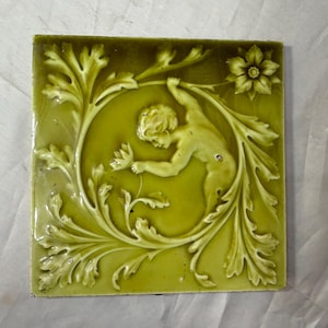 May include: A square, antique-style tile with a raised relief design. The tile features a central figure of a cherub reaching for a flower, surrounded by a circular frame of ornate leaves and floral elements. The tile is a muted green color.