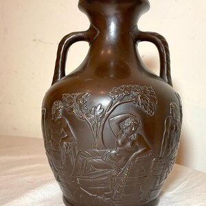 May include: A brown ceramic vase with two handles and a relief design of figures and trees. The vase has a dark brown glaze and a smooth finish.