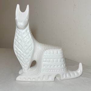 May include: White ceramic sculpture of a stylized animal with geometric patterns. The animal has a long, slender body with a long tail and a pointed head. The sculpture is in a sitting position.