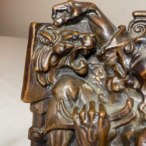 Puede incluir: Un par de sujetalibros de bronce que representan una escena de dos figuras, una con una cabeza de bestia que gru&ntilde;e, sosteniendo un libro. Los sujetalibros son detallados y tienen un aspecto r&uacute;stico y antiguo.