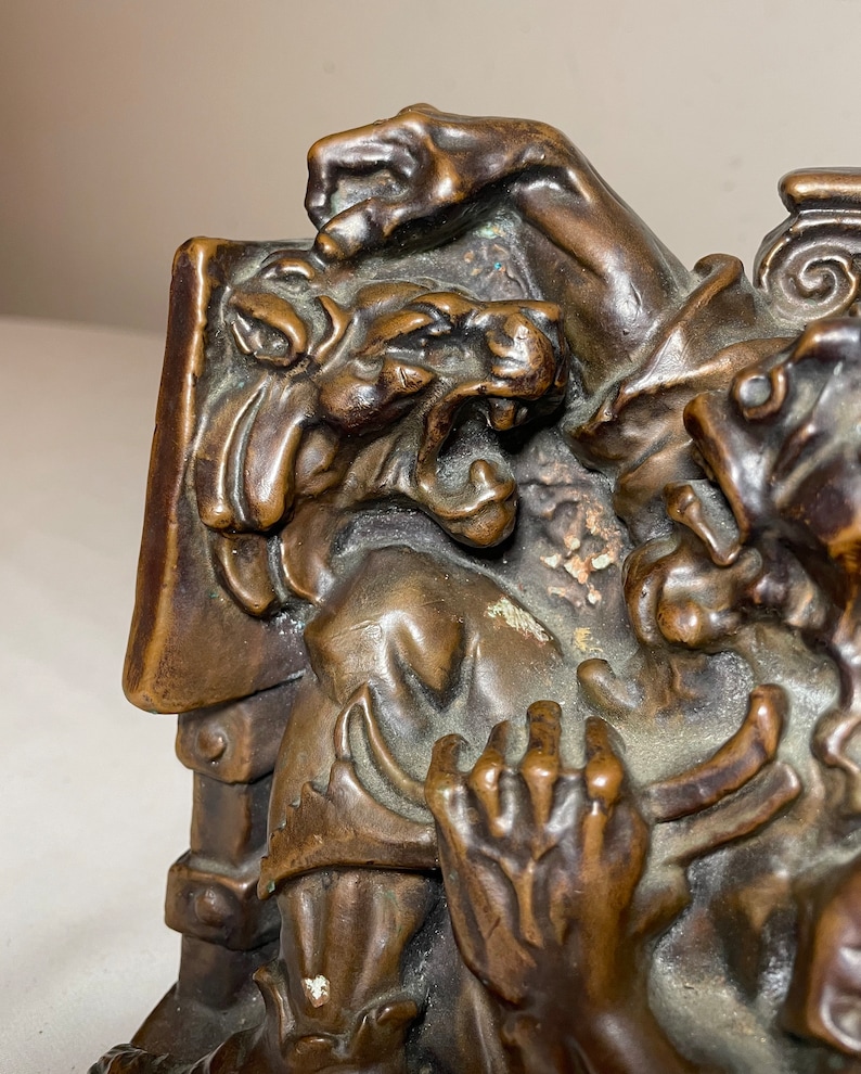 Puede incluir: Un par de sujetalibros de bronce que representan una escena de dos figuras, una con una cabeza de bestia que gru&ntilde;e, sosteniendo un libro. Los sujetalibros son detallados y tienen un aspecto r&uacute;stico y antiguo.