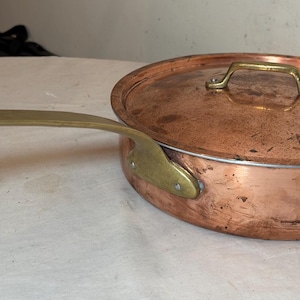 May include: A copper saucepan with a brass handle and lid. The lid has a brass handle. The pan is round and has a slightly worn appearance. The handle extends to the left.