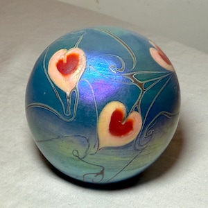 May include: A round, iridescent blue glass paperweight with a design of red and cream-colored hearts. The hearts are outlined with thin, dark lines, and the background has a swirling, organic pattern. The paperweight has a smooth, glossy finish.