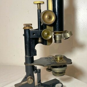 May include: An antique microscope with a black body and brass accents. The microscope has a vertical design with multiple adjustment knobs and lenses. The base is rectangular, and the overall appearance suggests a vintage scientific instrument.