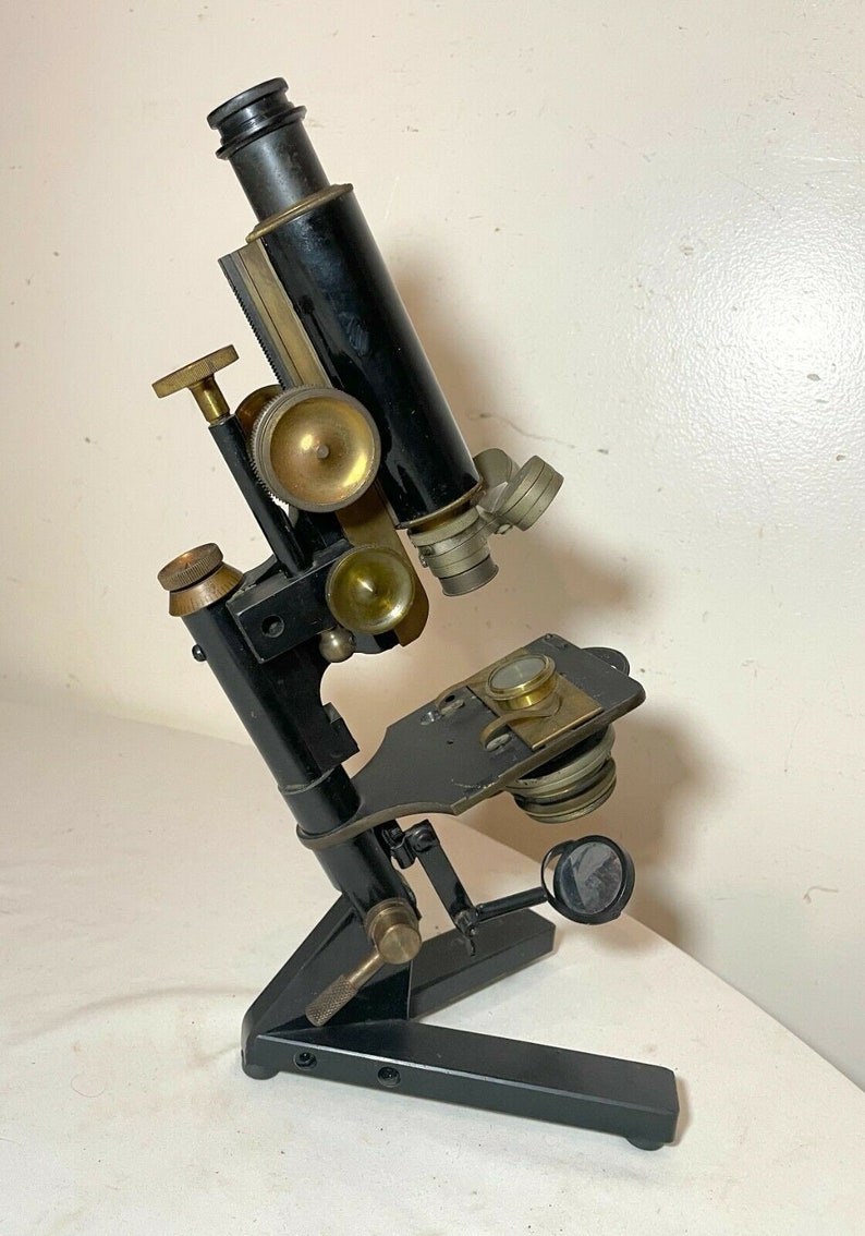 May include: An antique black microscope with brass accents. The microscope has a black base and a black tube with brass adjustment knobs and lenses. The microscope is angled towards the viewer and is set against a white background.