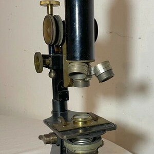 May include: An antique microscope with a black body and brass accents. It features adjustable knobs, lenses, and a stage for slides. The microscope is mounted on a tripod base, showcasing a classic scientific instrument.