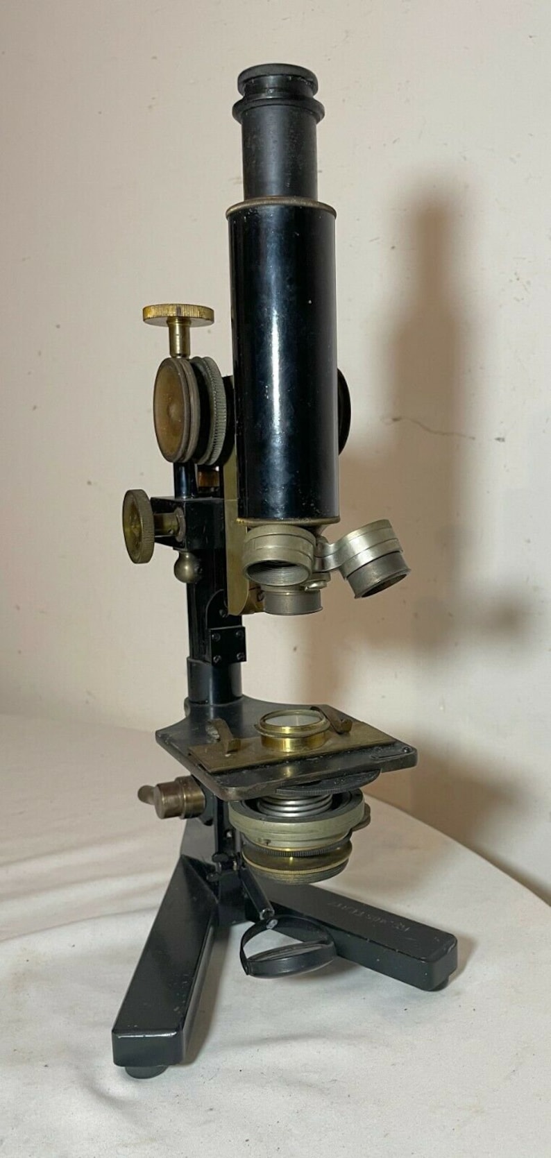May include: An antique microscope with a black body and brass accents. It features adjustable knobs, lenses, and a stage for slides. The microscope is mounted on a tripod base, showcasing a classic scientific instrument.
