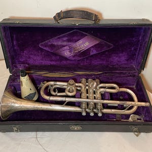 May include: A silver trumpet with engraved details in a black velvet-lined case. The case has a brass latch and a leather handle. The inside of the case has a purple velvet lining with a label that reads "C.G. Conn Ltd. Elkhart, Indiana, U.S.A."