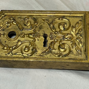 May include: Ornate, antique brass door plate with a keyhole and decorative floral and scrollwork design. The rectangular plate has a rounded edge on the left side and a vertical floral border on the right.