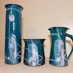 May include: A set of three teal ceramic vases with abstract white, pink, and blue painted designs. The set includes a tall cylindrical vase, a shorter vase, and a pitcher with a handle.