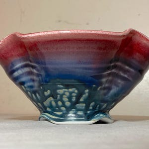 May include: A small, ceramic bowl with a ruffled rim. The bowl is glazed in shades of pink, blue, and green, with a white speckled design.